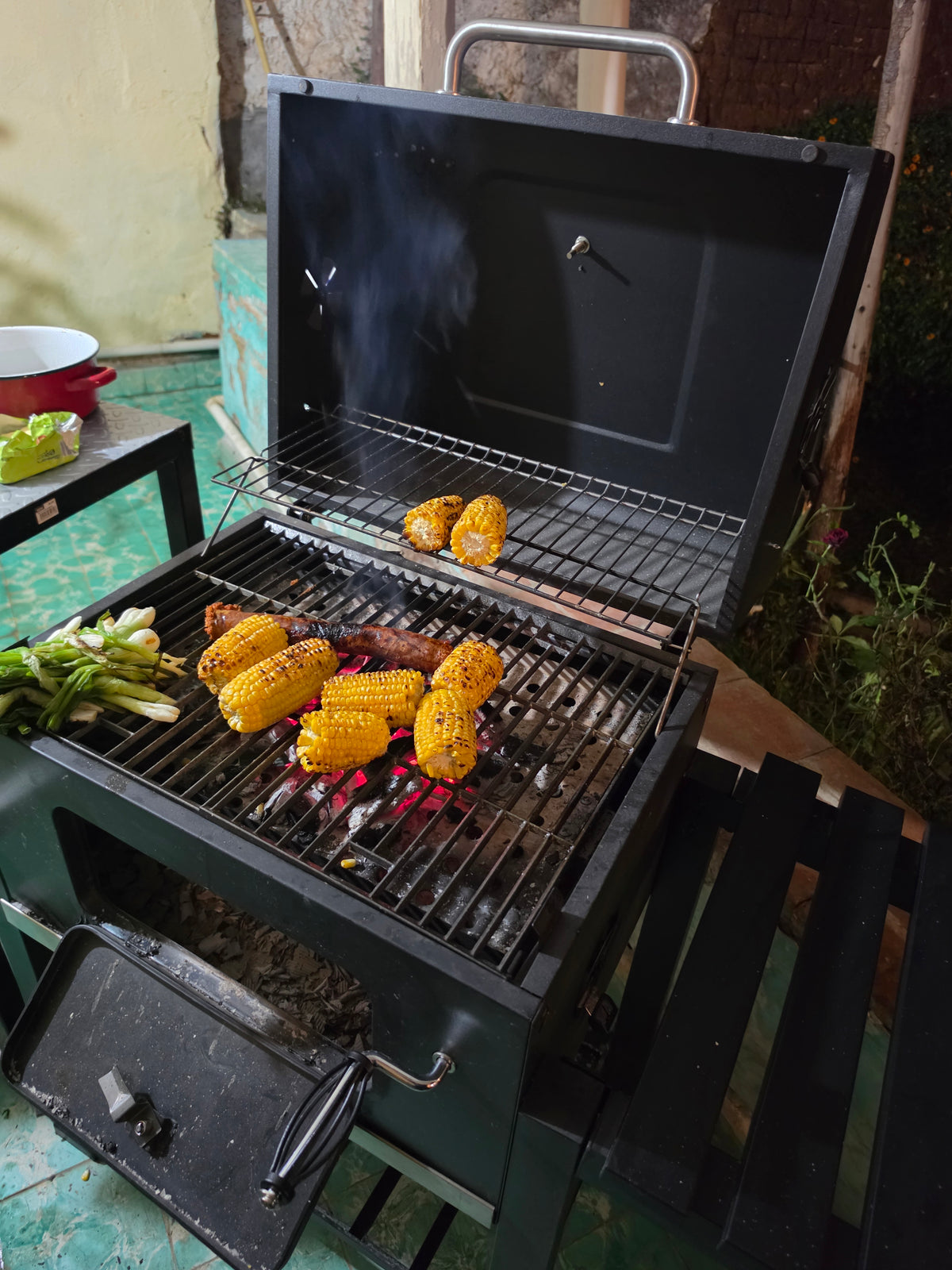 Asador de carbón Fiero con la tapa abierta asando elotes, cebollitas y carne, con rejilla superior